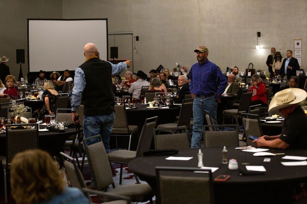 A presenter points to a projector screen in a ballroom filled with round tables, as an attendee walks toward the front.