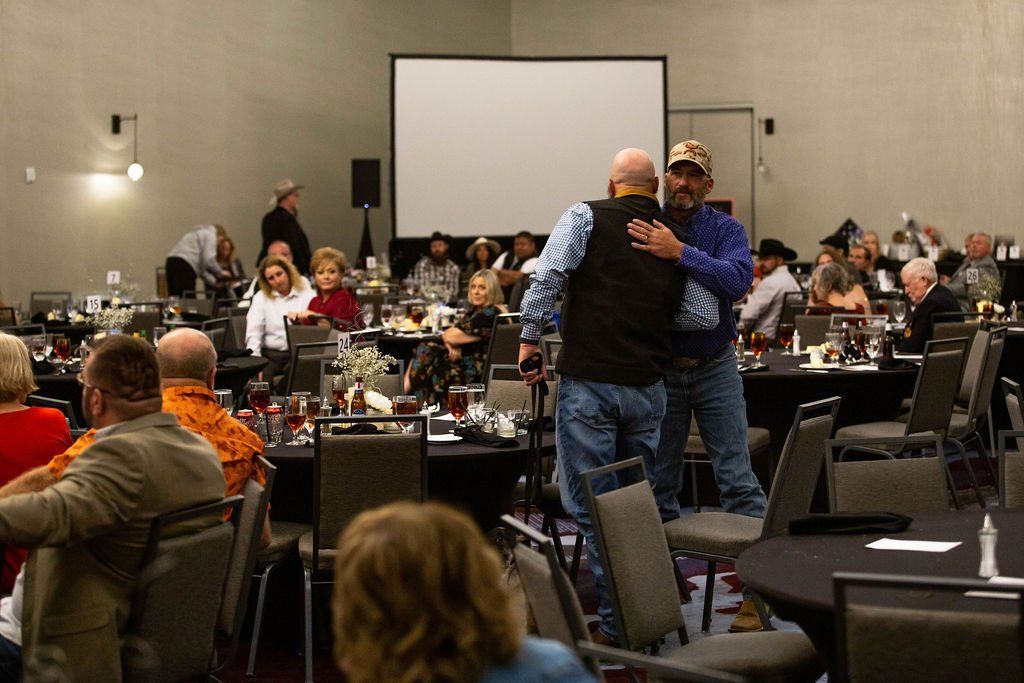 Two people embrace in a banquet hall while attendees sit at tables in the background.