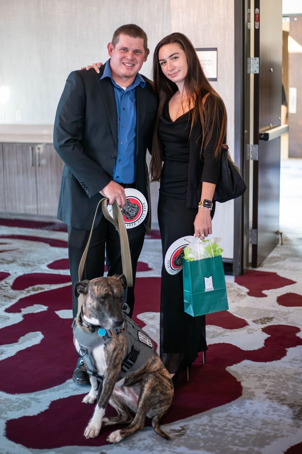 A person in a suit and another in a black dress stand with a service dog wearing a gray vest on a patterned carpet.