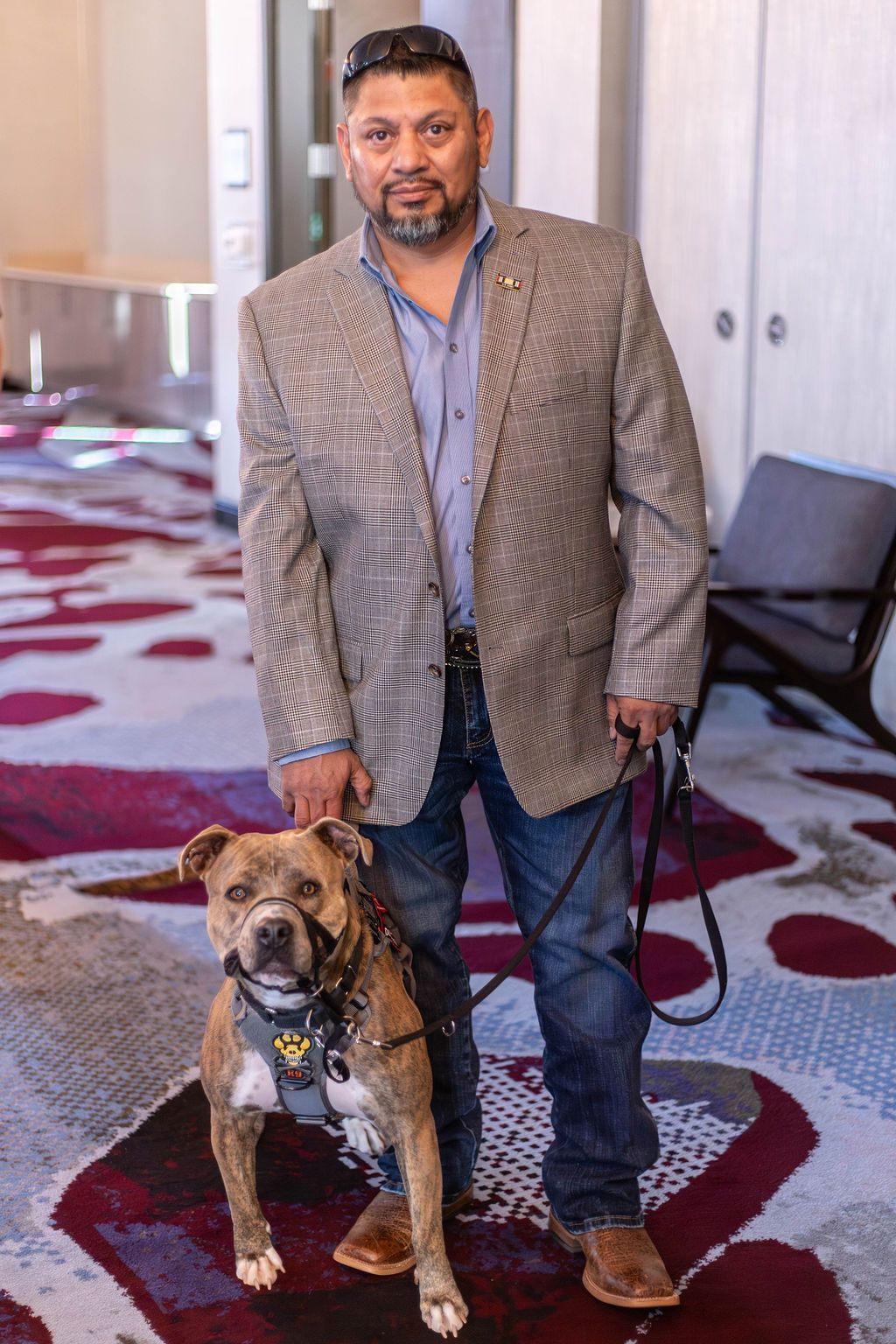 A man in a blazer and jeans stands with a brindle service dog wearing a gray vest on a patterned carpet.