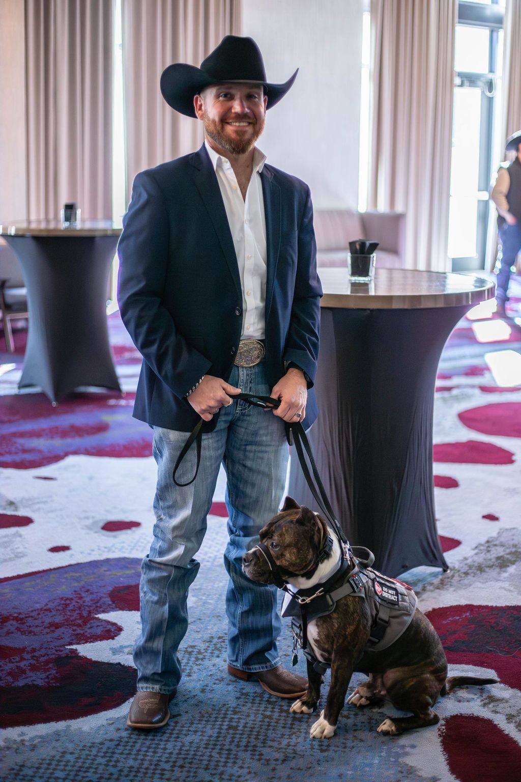 A man in a cowboy hat, suit jacket, and jeans stands indoors holding a leash attached to a brindle service dog.