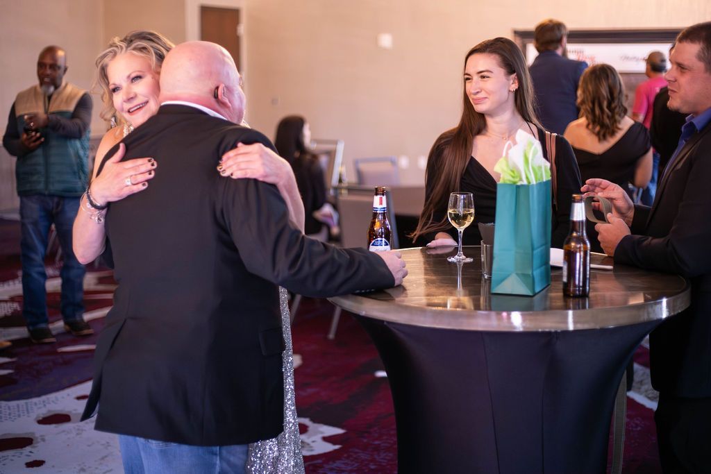 Guests gather at a table with drinks and gift bags in a banquet hall, while two people embrace in the foreground.