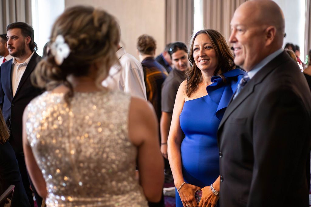 People in formal attire talk at an indoor event; one person wears a silver sequined dress, another a blue one-shoulder dress.