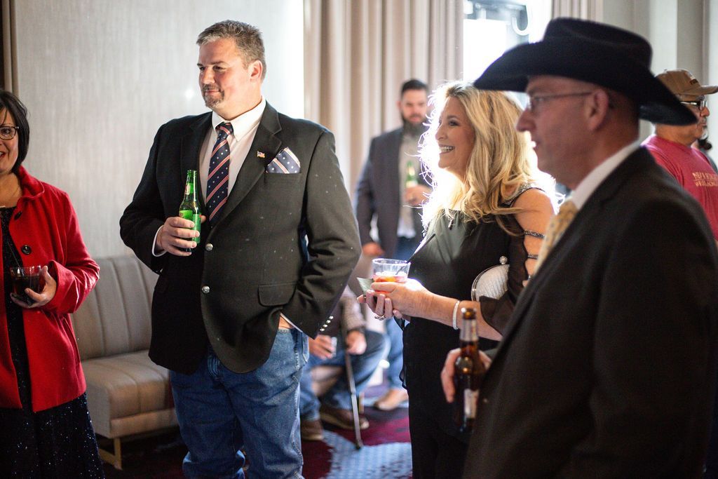 A group of people dressed in formal attire socializes in a brightly lit indoor space, holding drinks and chatting.