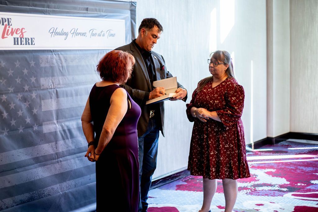 A person in a suit presents a small, open box to another person in a dark dress, standing before a flag-themed backdrop.