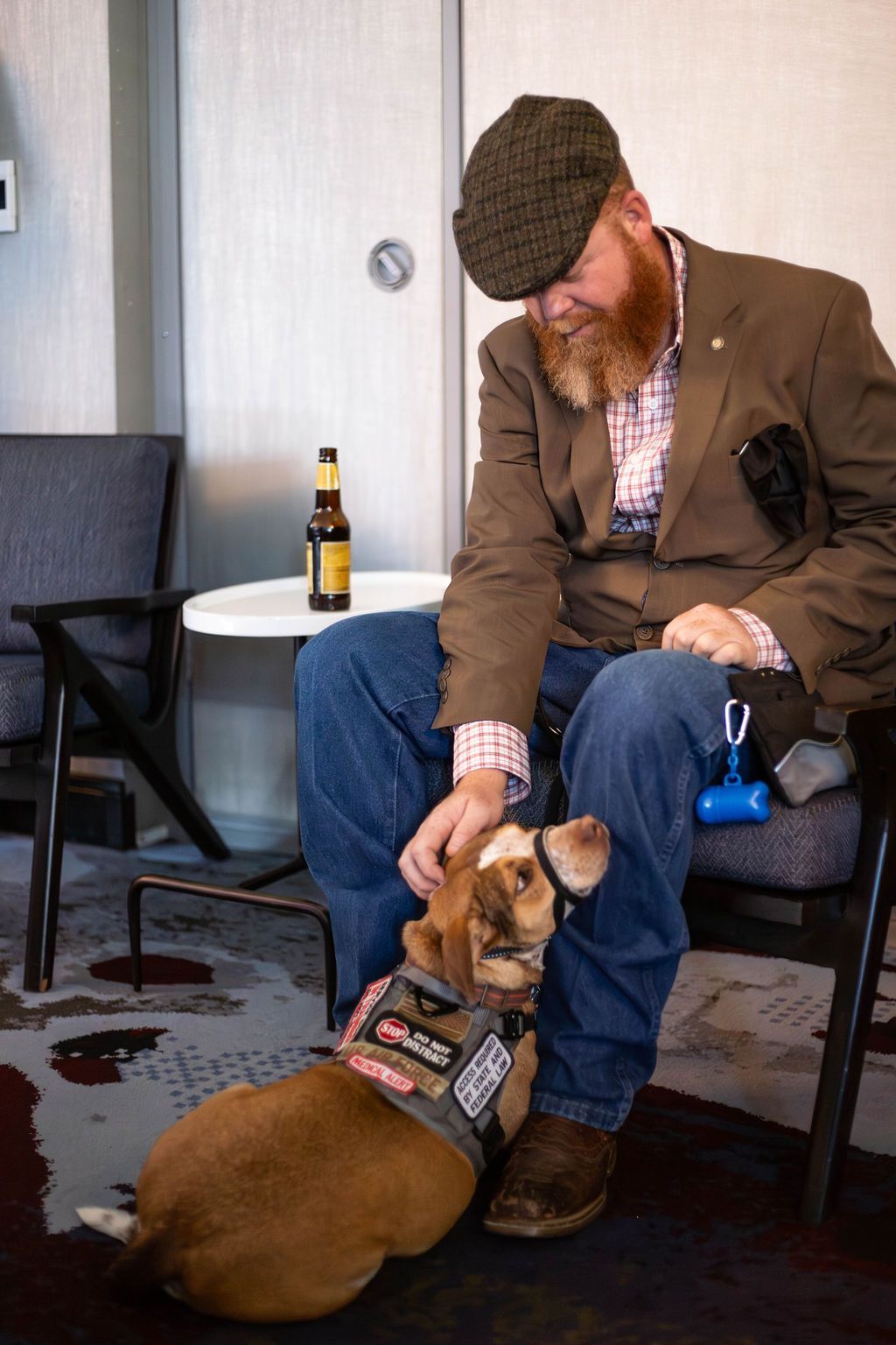 A bearded person in a brown blazer and cap pets a small, harnessed dog sitting between their legs in a room.