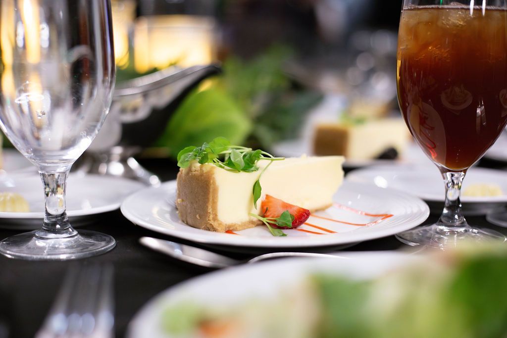 A slice of cheesecake garnished with a strawberry and greens on a white plate, flanked by a wine glass and iced tea.