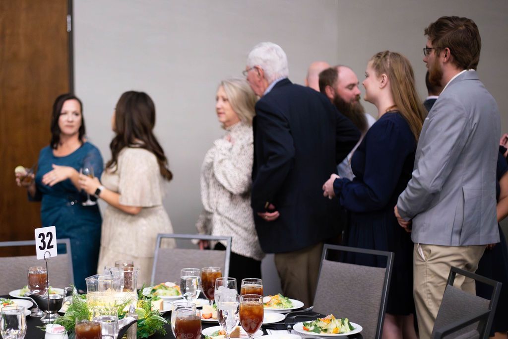 A group of people standing in a line at a catered event, gathered near tables with plates of food and drinks.