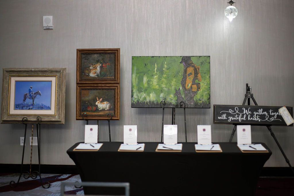 A display table with four framed paintings and a sign on a wall, with auction bid sheets underneath.