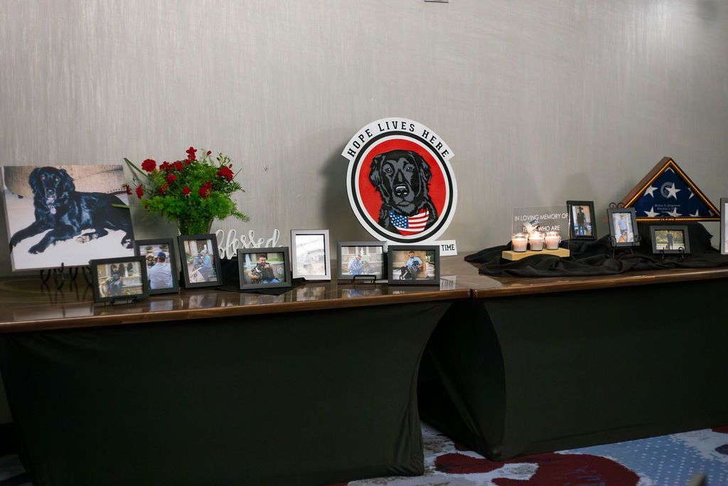 A memorial table featuring photos, red roses, a black dog emblem, and a folded American flag on dark green draped tables.