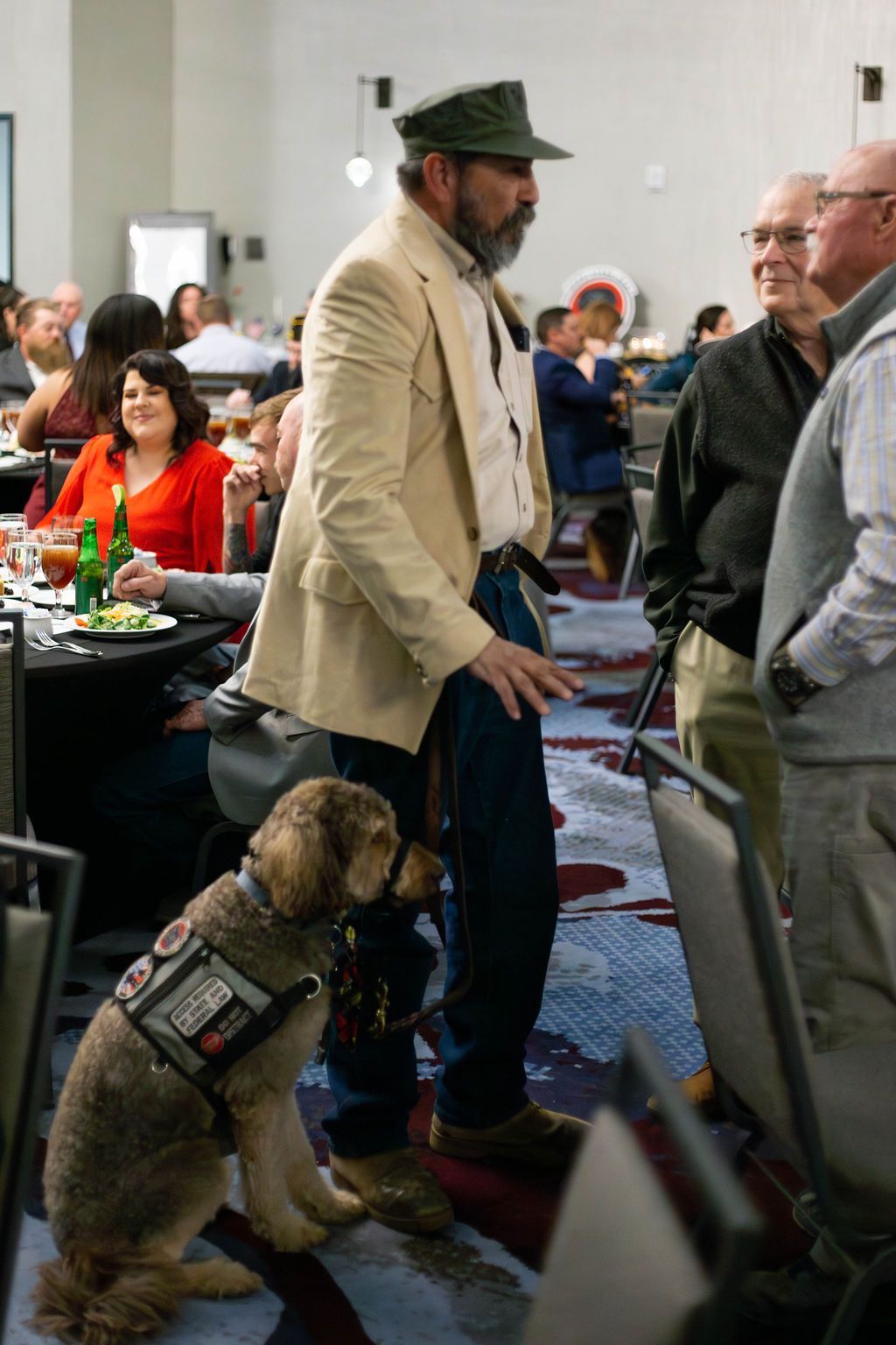 A man in a beige jacket and hat speaks to two men, with a service dog wearing a vest sitting attentively at his side.