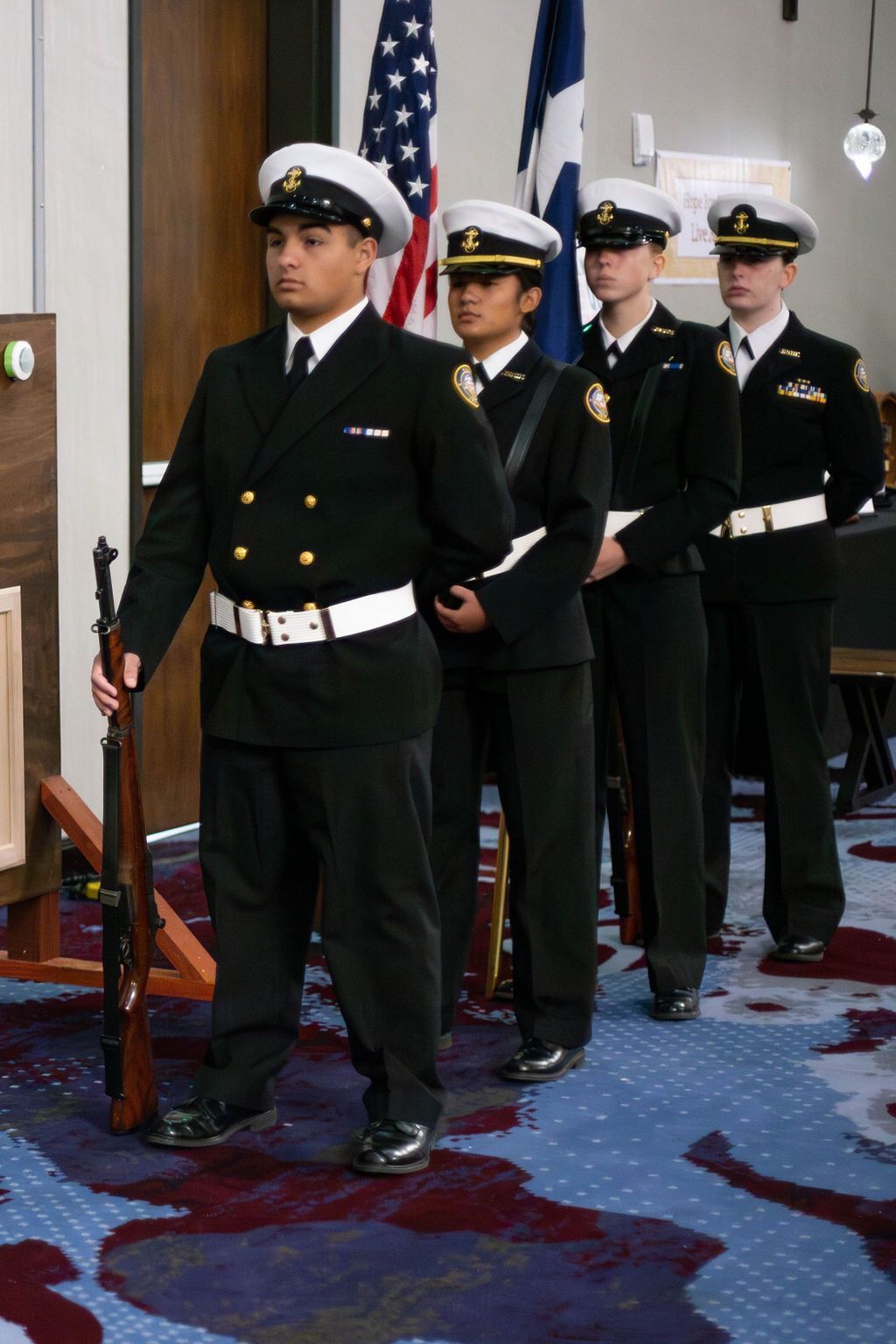 Four members of a color guard in dark naval-style dress uniforms standing in a line at attention with rifles.