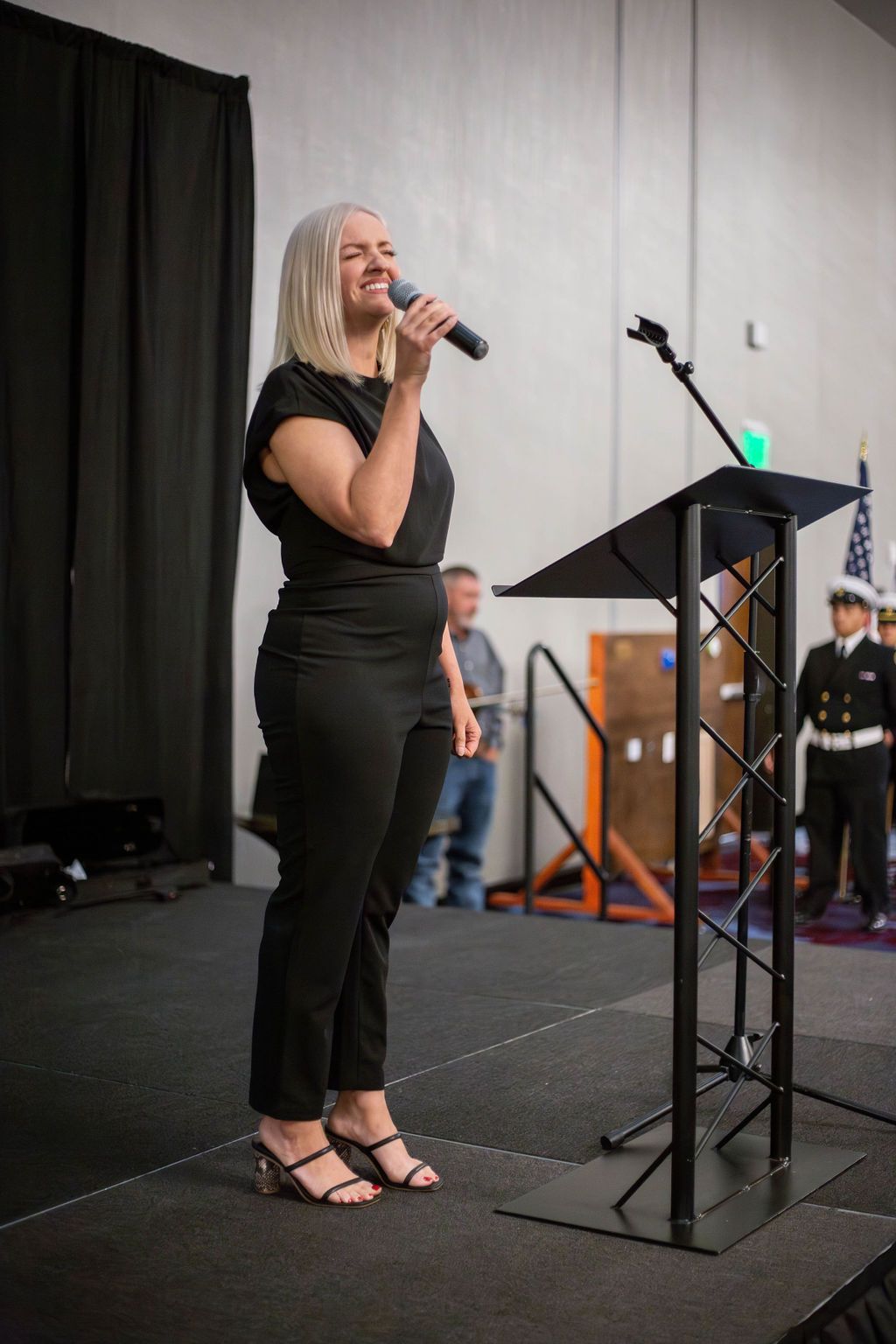A person in a black outfit stands at a podium on a stage speaking into a microphone, with a person in uniform visible nearby.
