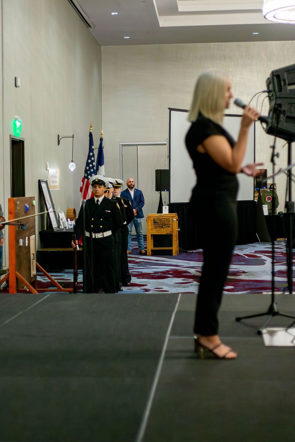 A person speaks into a microphone on a stage while members of a military color guard stand at attention in the background.