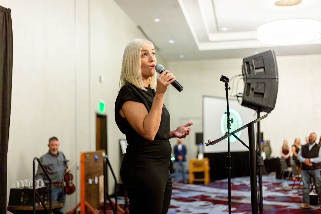 A person in a black outfit speaks into a microphone on stage, with a musician holding a violin standing in the background.