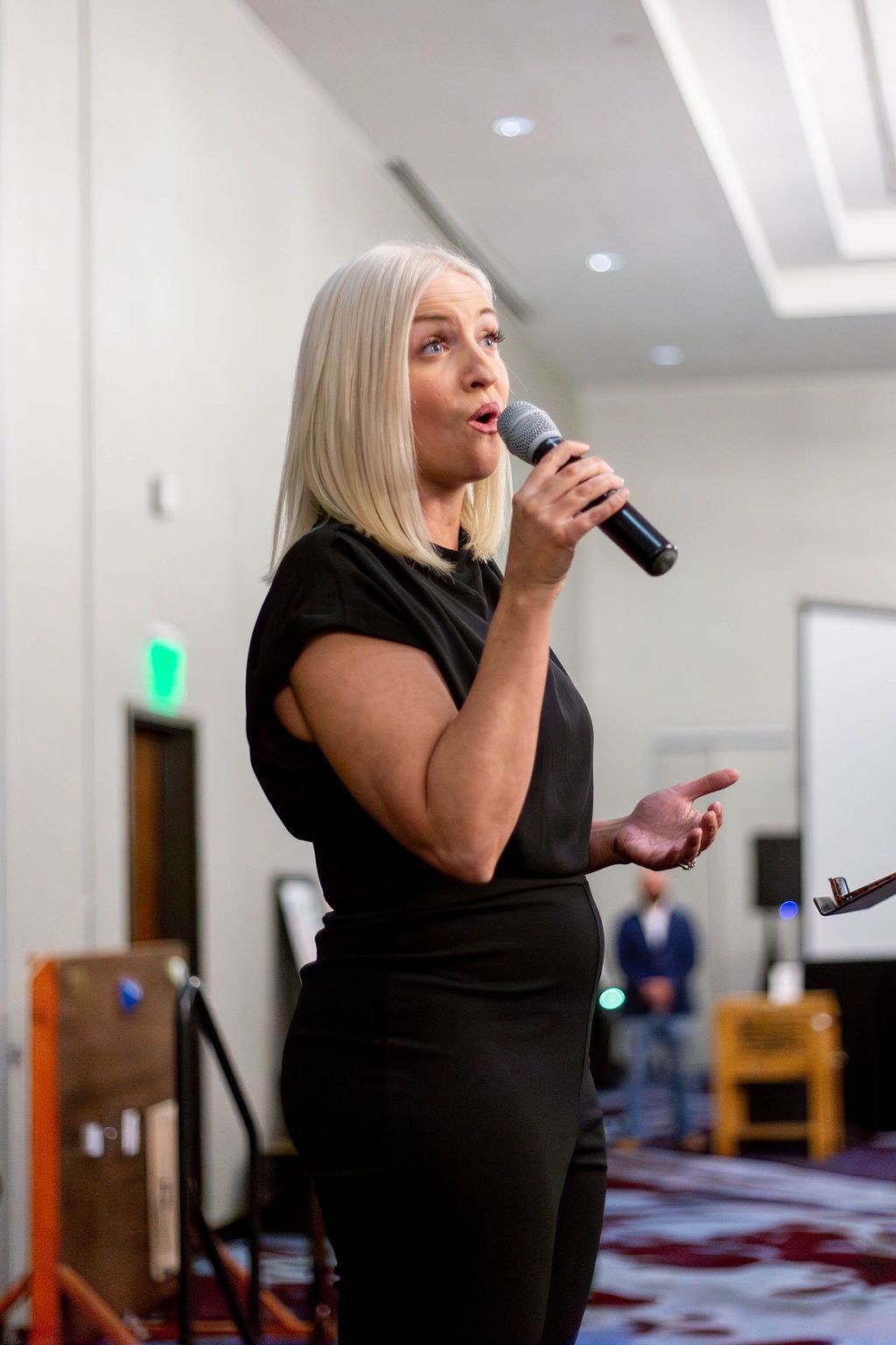 A person with short blonde hair in a black top speaks into a microphone during a presentation in an indoor event space.
