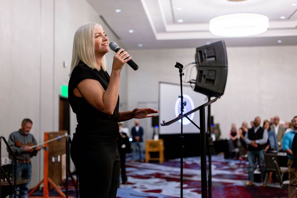 A blonde person speaks into a microphone at a podium in a conference hall with an audience in the background.