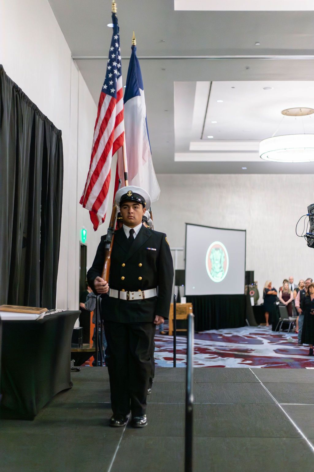 A member of a color guard in a dress uniform marches on a stage holding the U.S. and Texas flags in a convention hall.