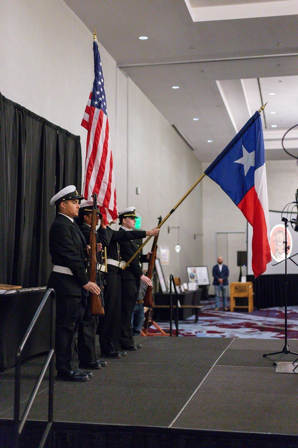 A color guard stands on a stage holding the U.S. and Texas flags and ceremonial rifles in a formal indoor setting.