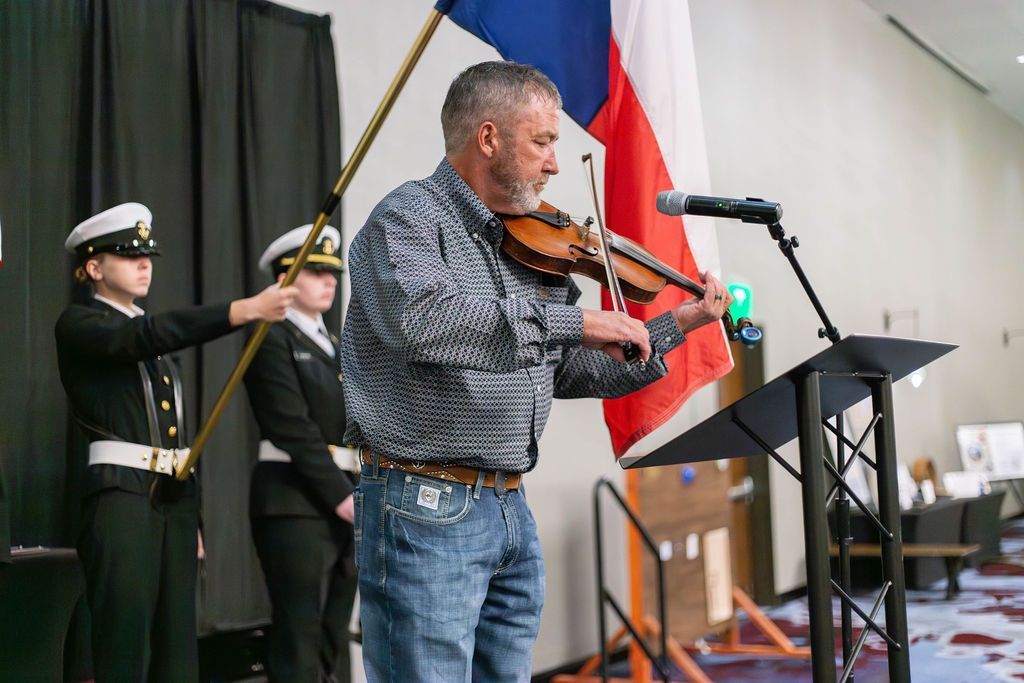 A man in a patterned shirt plays a violin at a podium with two uniformed color guard members standing behind him.