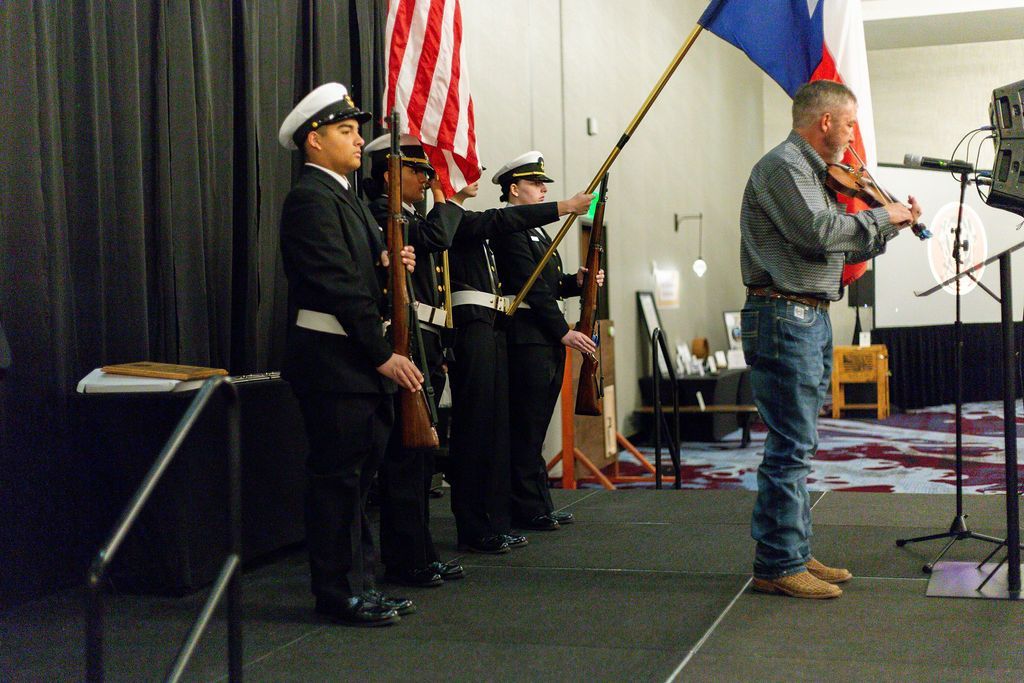 A color guard stands at attention holding flags and rifles while a person performs on a violin on a stage.