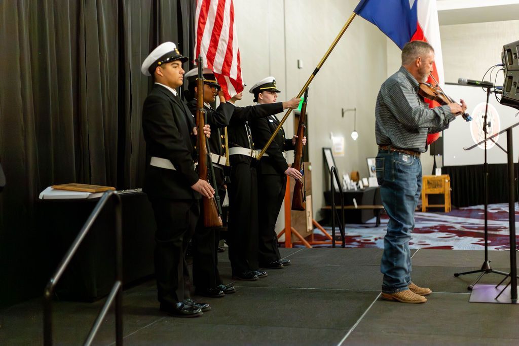 A violinist plays on stage next to an honor guard holding American and Texas flags on a stage with a black curtain.