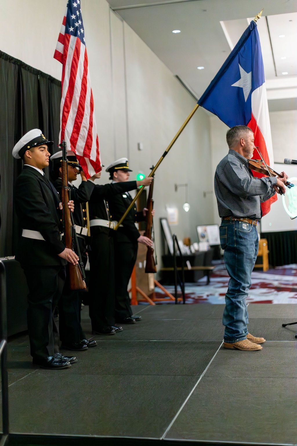 A color guard stands on a stage holding US and Texas flags while a man plays a violin.