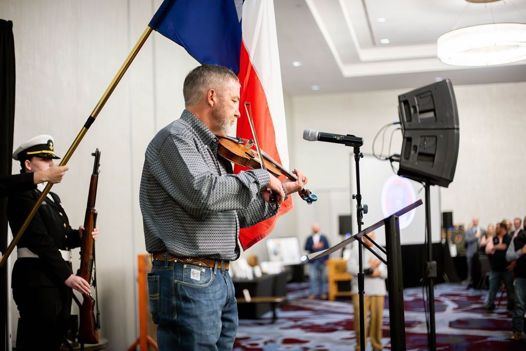 A man plays the violin at an indoor event, standing before a Texas flag while a person in a uniform holds a rifle.