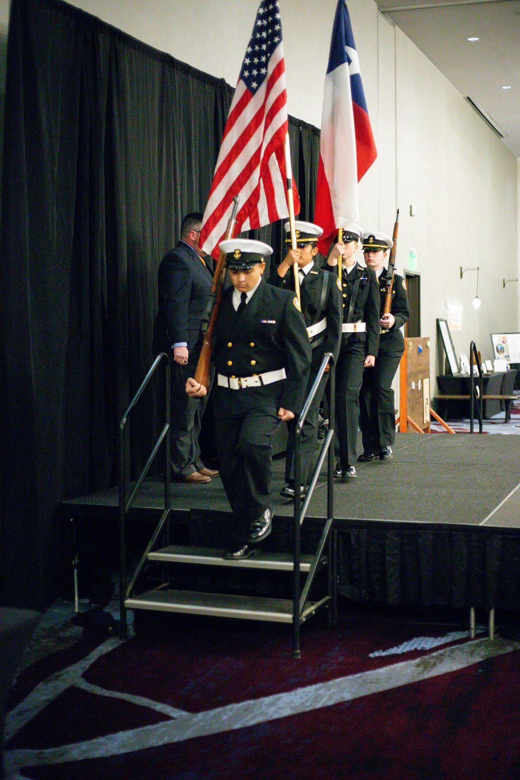 A color guard in navy uniforms marches down stage steps carrying the US and Texas flags, with a person standing nearby.