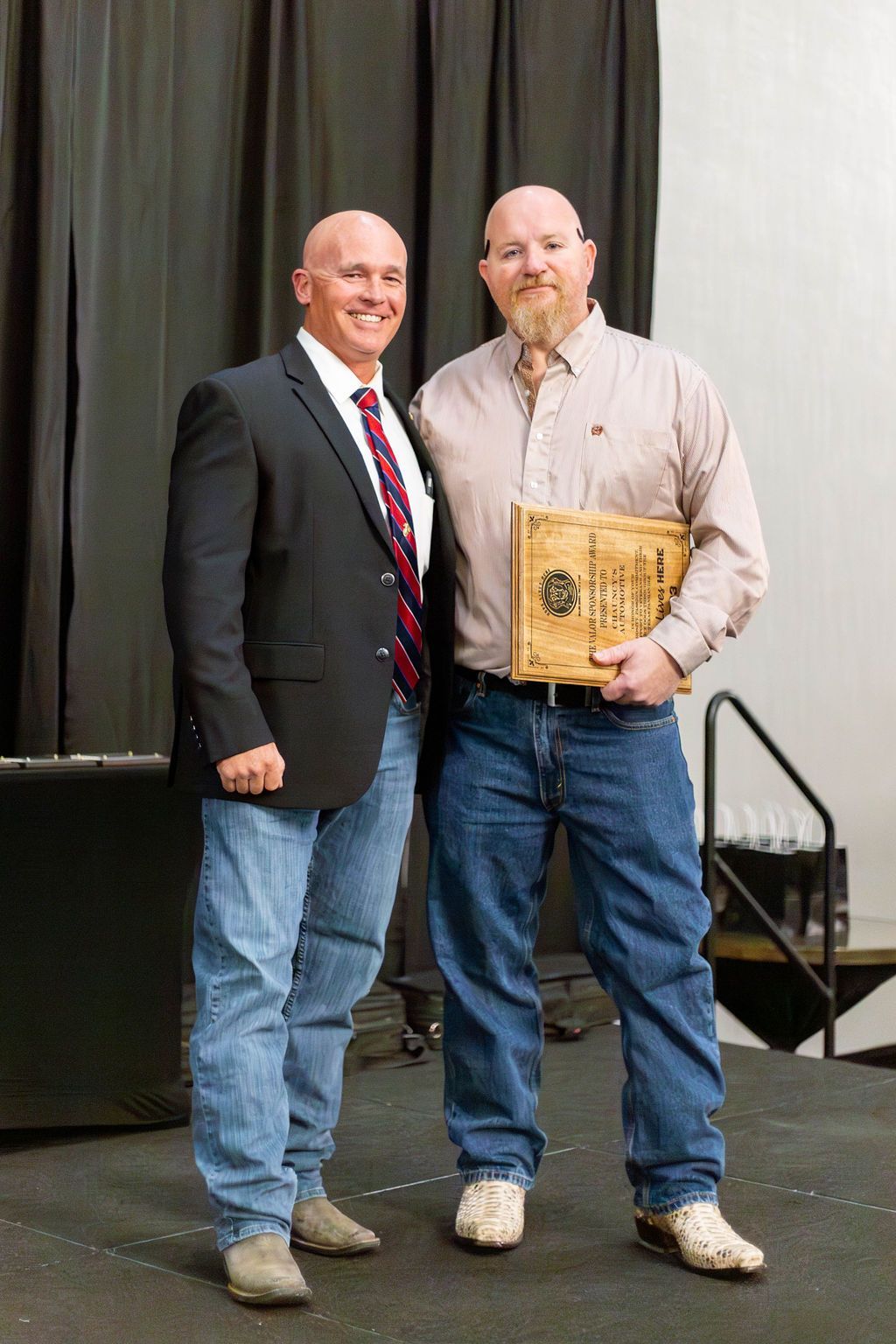 Two men smiling while standing on a stage, one in a suit and tie, the other in a button-down shirt holding an award.