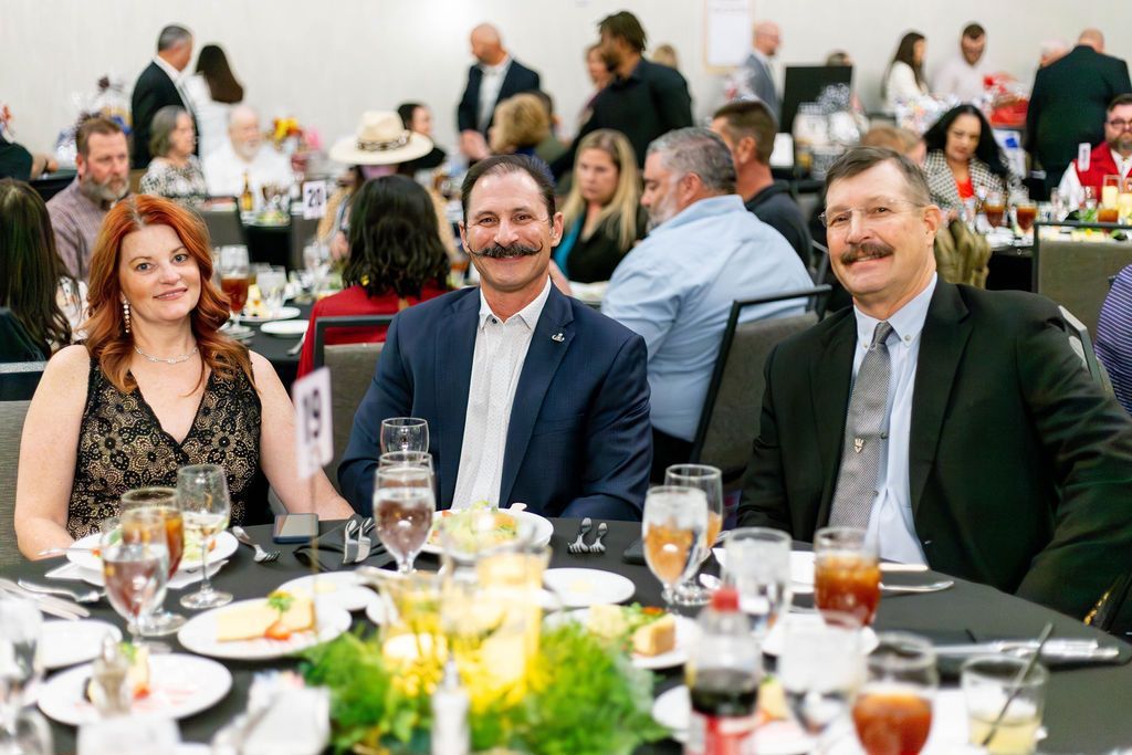 A trio of formally dressed people sit at a round, black-clothed table at an event, smiling toward the camera.