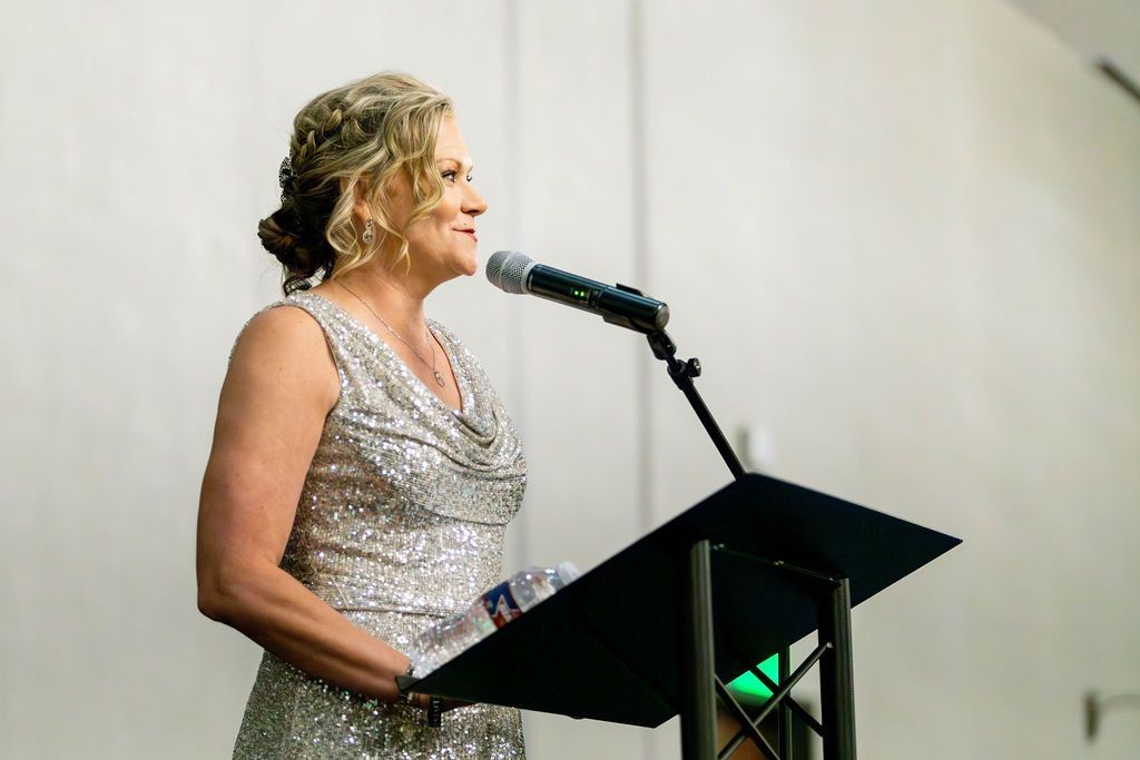 A person in a shimmering silver dress stands at a podium, speaking into a microphone in a brightly lit, simple room.