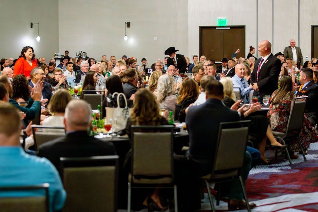 A large, formal audience in a banquet hall applauds as a speaker addresses the room from the front of the tables.