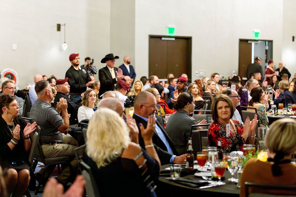 People in formal attire applauding in a brightly lit banquet hall, some standing and others seated at round tables.