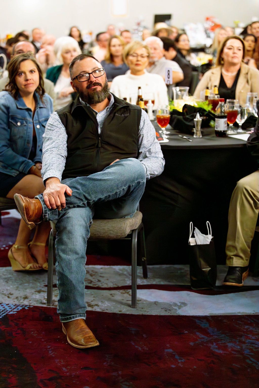 A man with a beard sits at a table in a crowded event hall, wearing a vest and jeans, with attendees seated nearby.