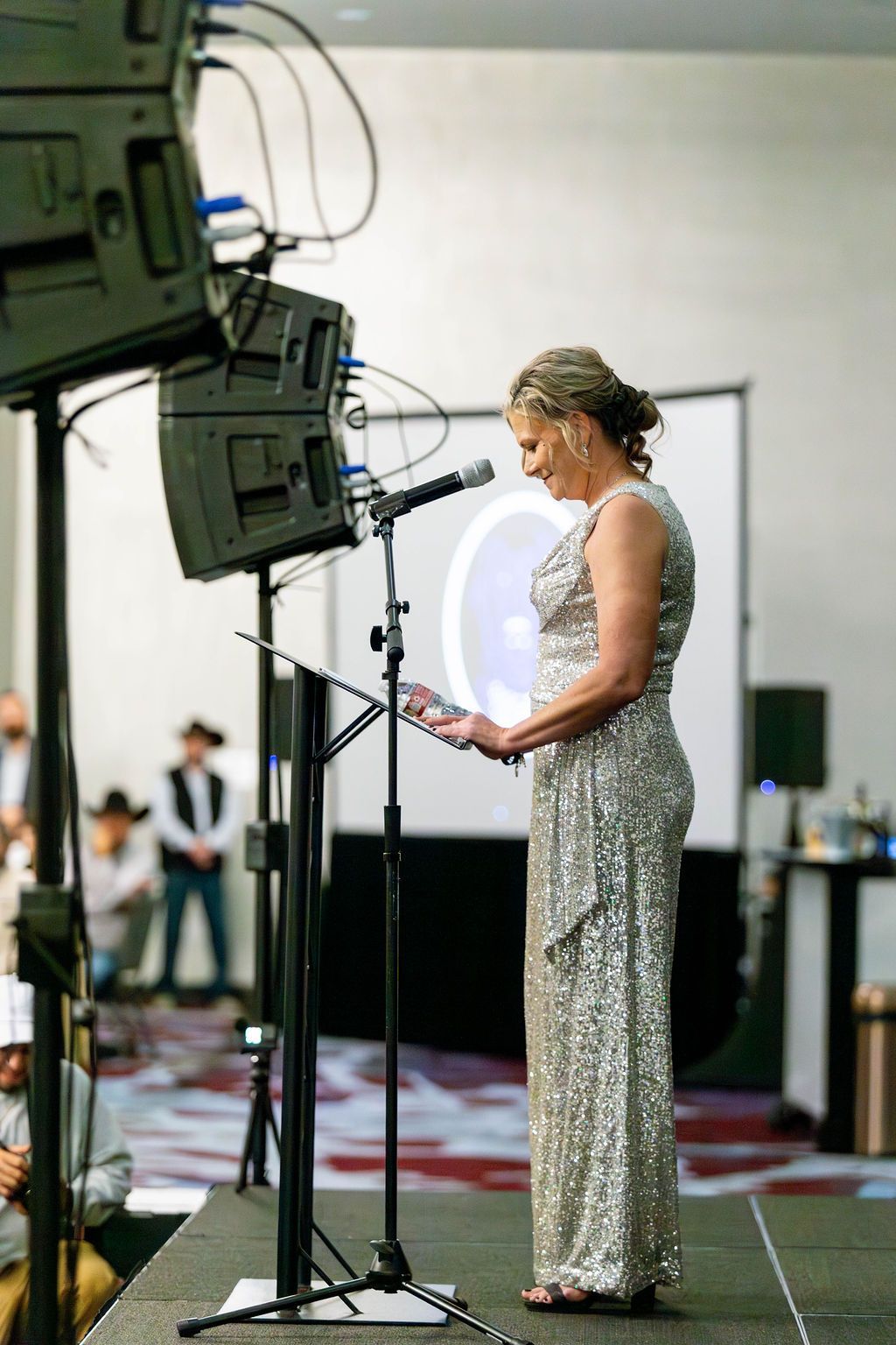 A person in a shimmering silver gown stands at a podium, speaking into a microphone at an indoor event.