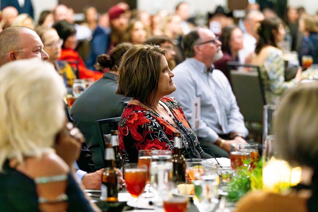 People sit at tables in a banquet hall, listening to a presentation during a formal event.
