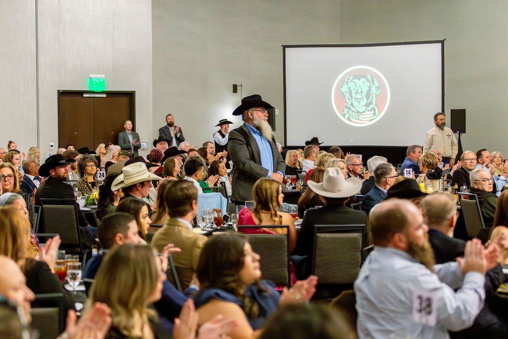 A speaker in a cowboy hat stands before an audience in a conference room, with many people applauding toward a large screen.