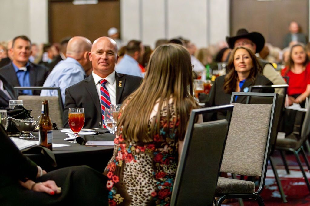 Attendees sit at round tables in a conference hall, listening to speakers during a formal event.