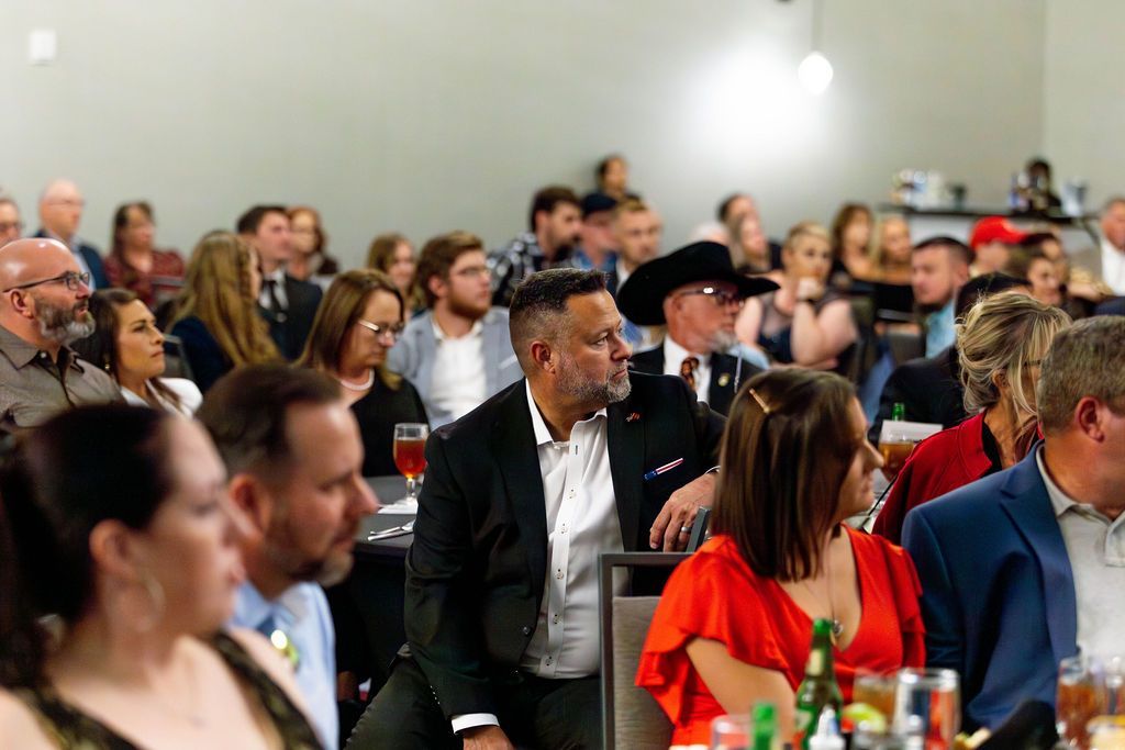 A diverse audience listens attentively at an indoor event, with people seated at tables in a large, bright hall.