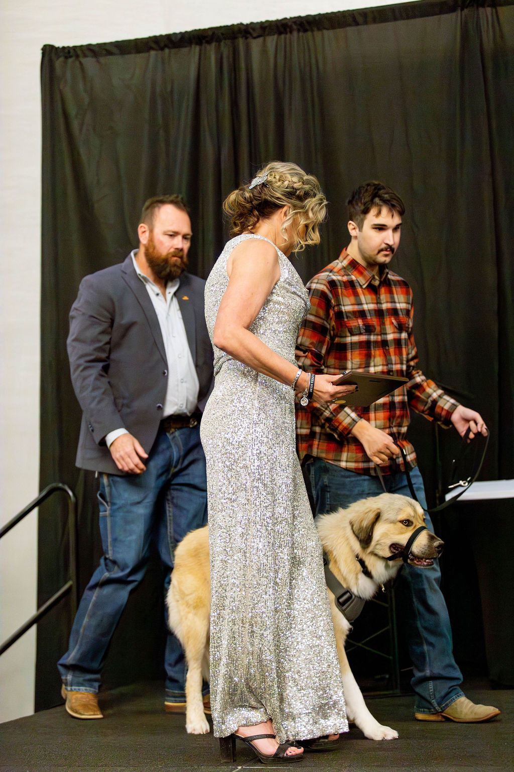 Two people in professional attire stand on stage with a large, light-colored dog on a leash in front of a black curtain.