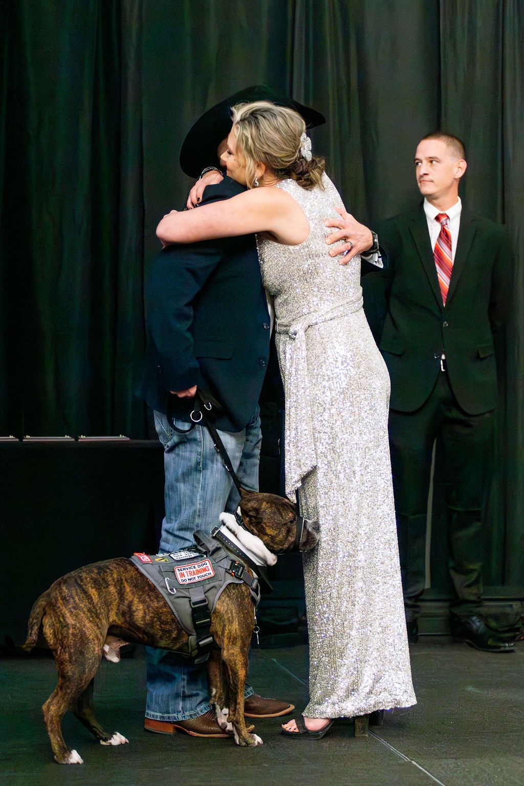 A person in a cowboy hat and a person in a sequined gown embrace as a service dog on a leash stands between them.
