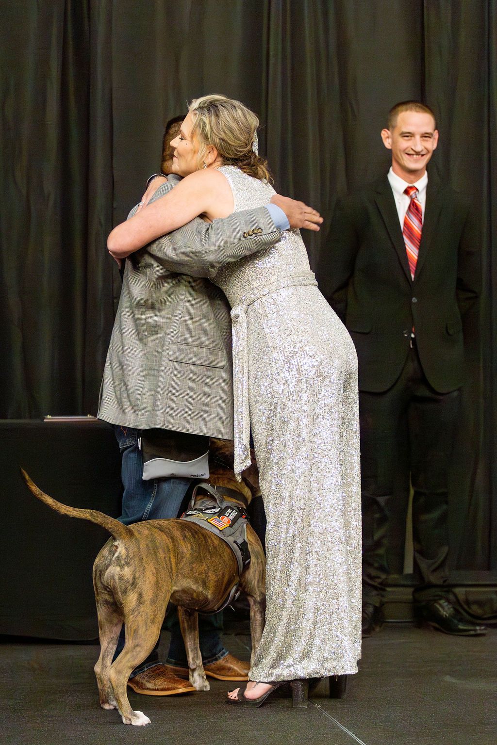 A person in a shimmering gown hugs another person, both standing on a stage with a brindle service dog and a smiling man.