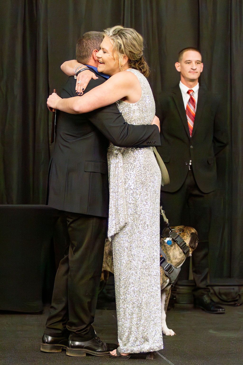 A woman in a sparkly silver dress hugs a person in a black suit. A dog in a harness and a person in a suit stand nearby.