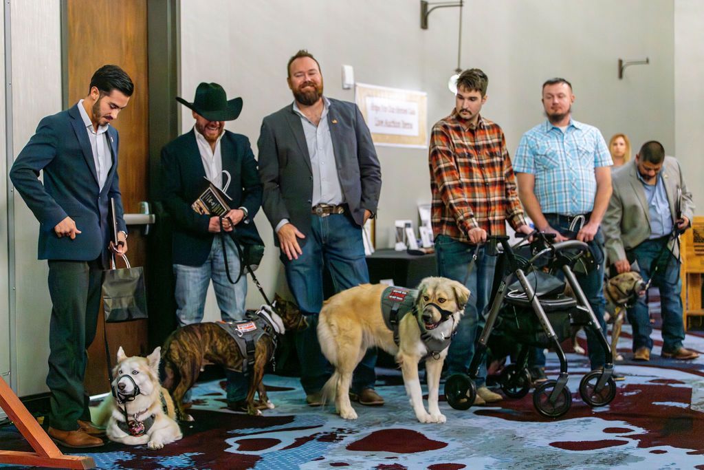 A group of people standing indoors with service dogs, including a brindle dog, a light-colored dog, and a stroller.