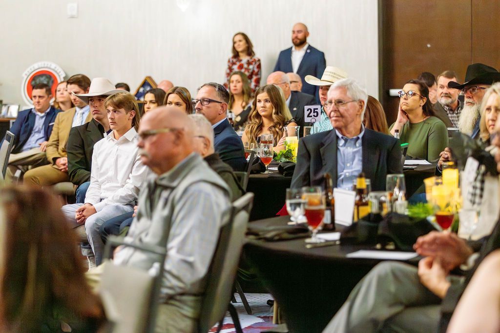 Attendees sit at tables in a brightly lit banquet hall, listening to a presentation during an indoor event.