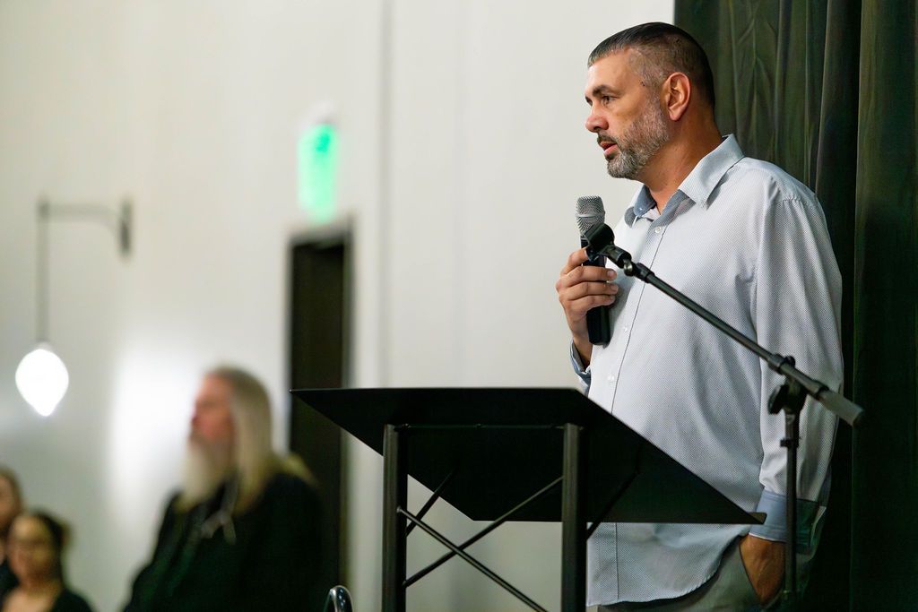 A person speaks into a microphone at a podium in a brightly lit room. A blurred person sits in the background.