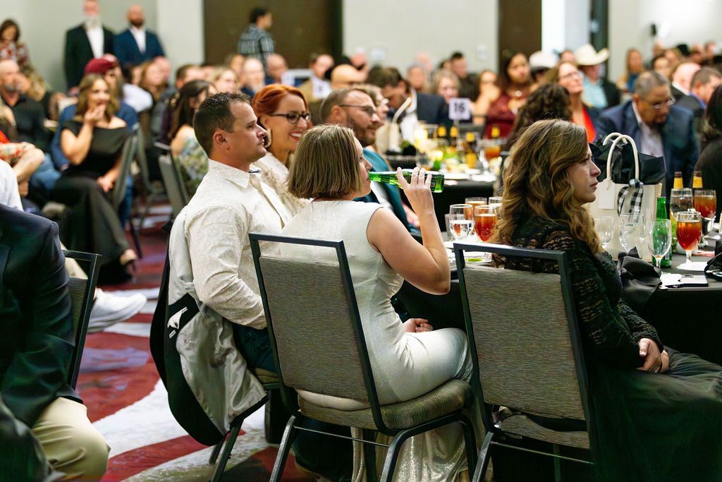 People sit at tables during a banquet in a large room, with one person in the foreground gesturing with a drink in hand.