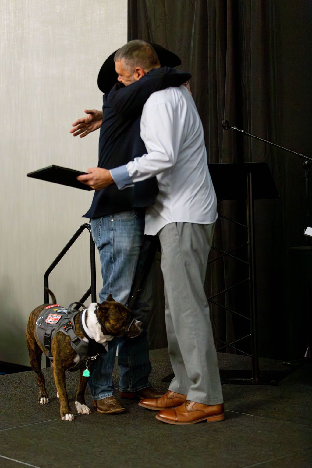 Two people embrace on a stage, with one wearing a hat and the other a white shirt, next to a service dog in a harness.