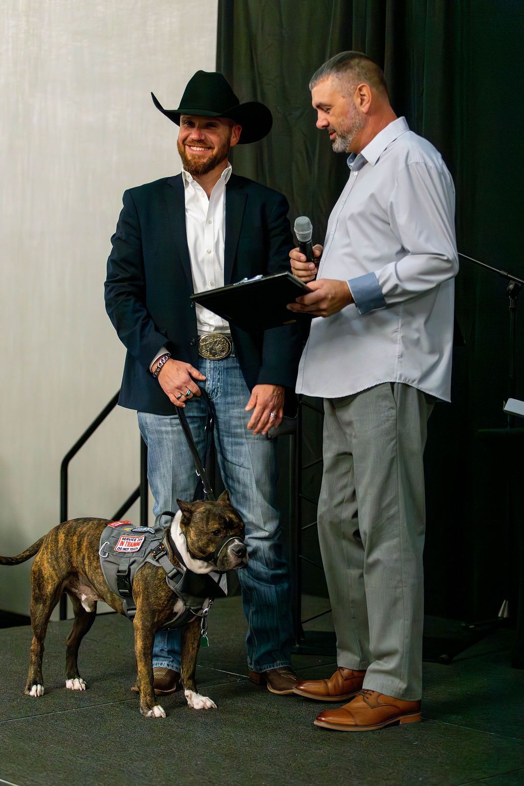 A person in a cowboy hat and suit stands with a brindle service dog while another person speaks into a microphone nearby.